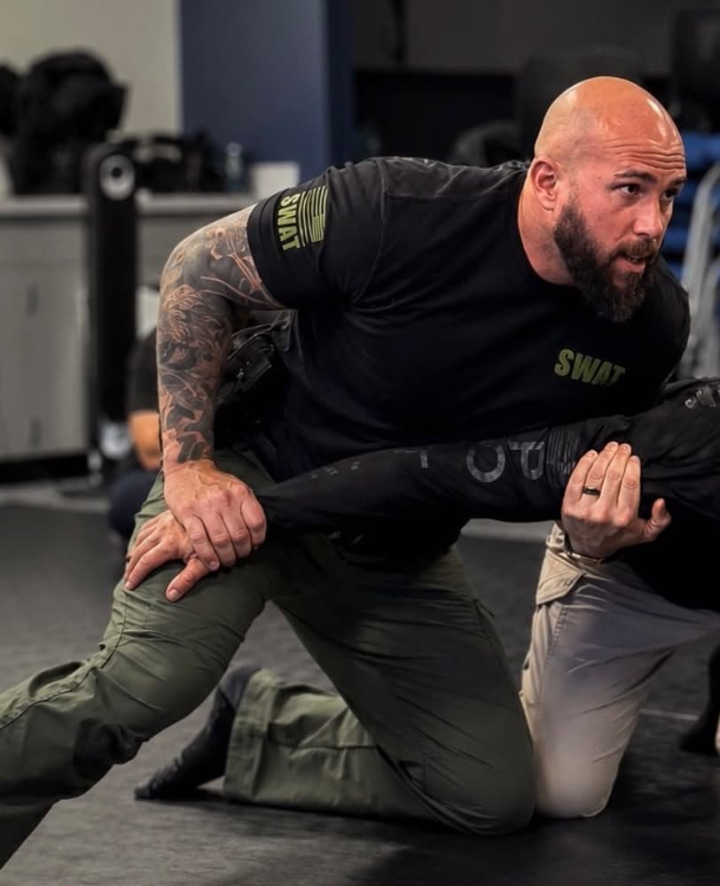 Hector on the mats, kneeling and controlling a training partner's arm